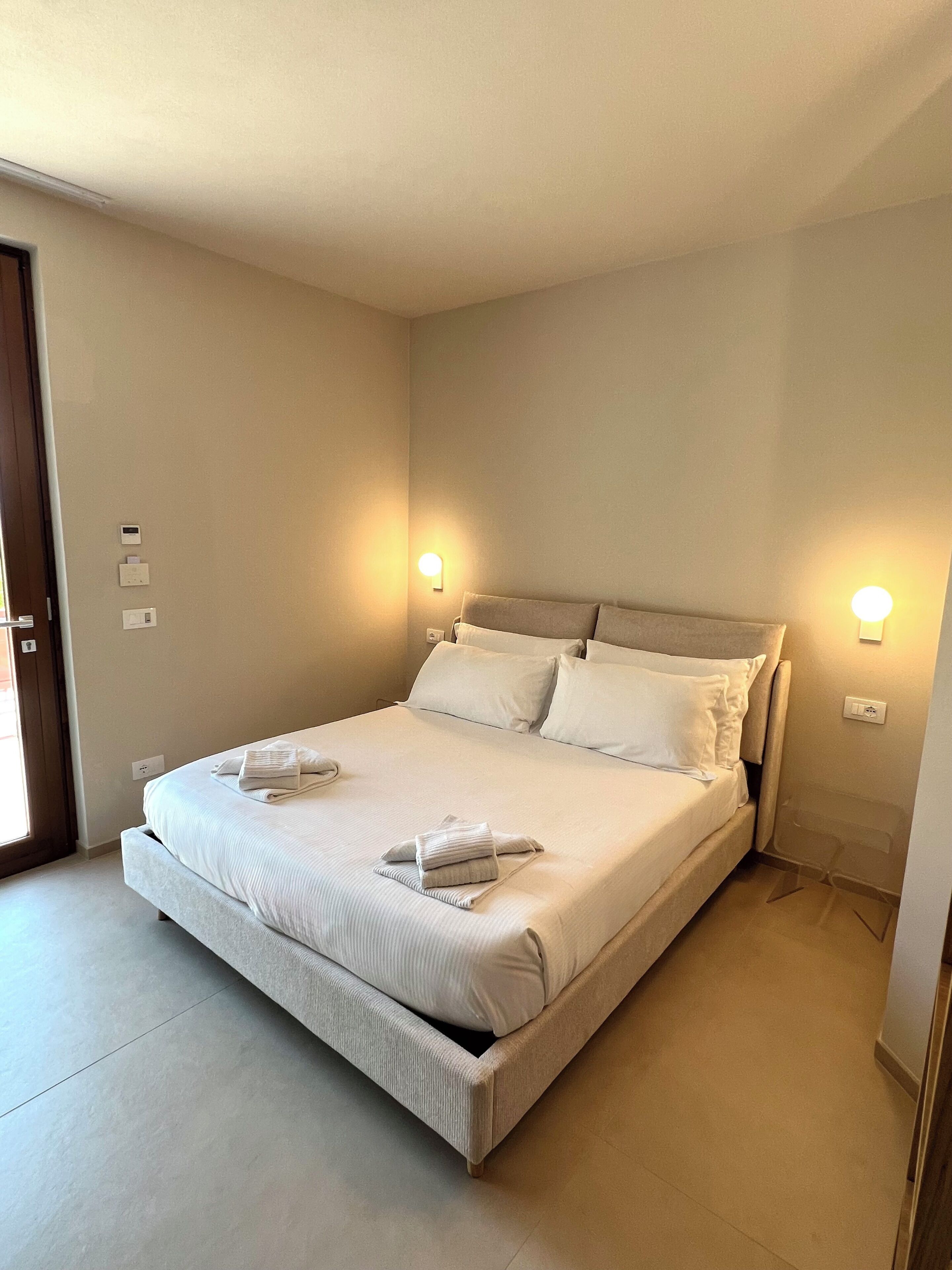 Un lit bien fait, avec des oreillers et une serviette pliée, une table de chevet avec une lampe, et une porte sur la gauche.