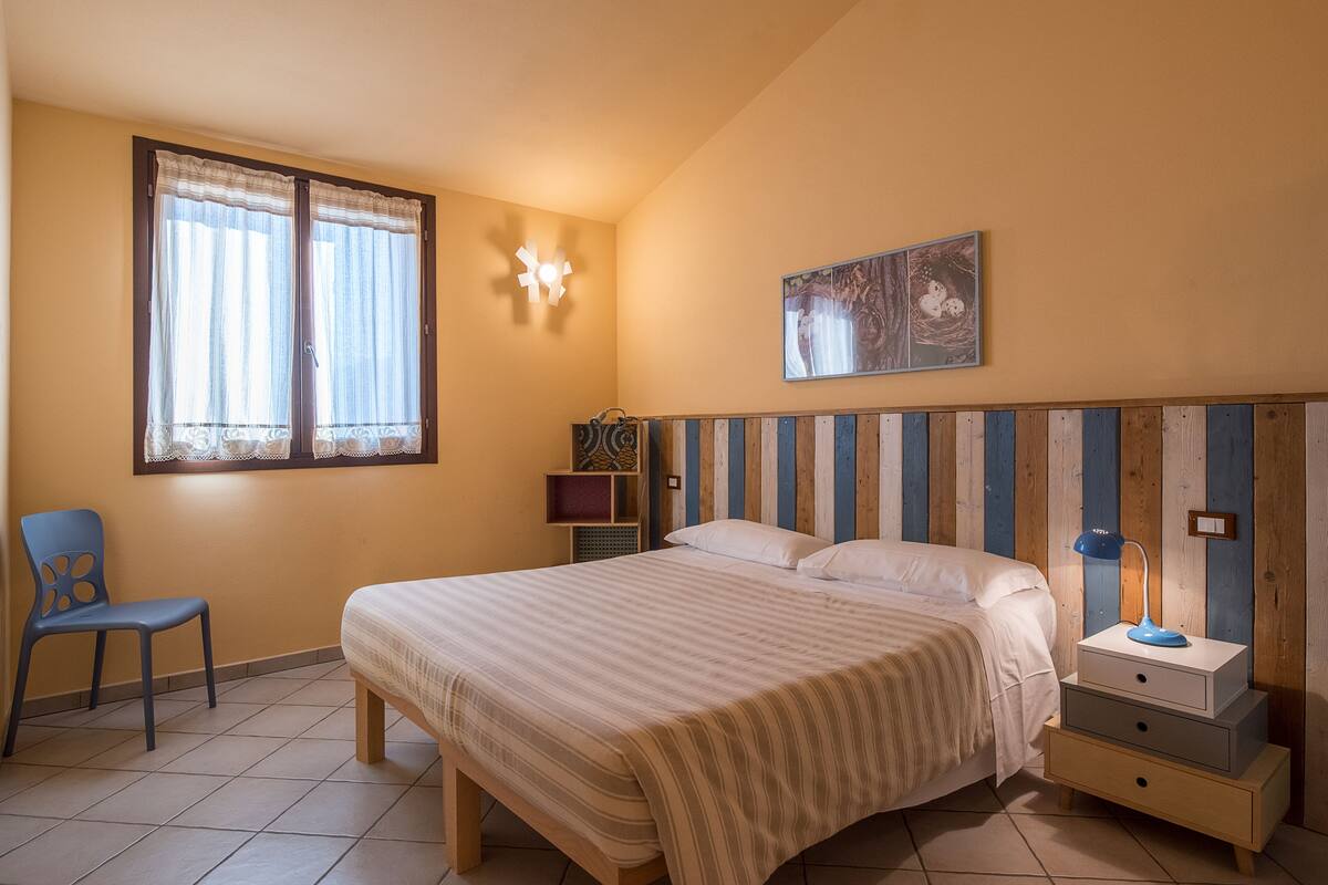 Un dormitorio con una cama, una silla, una mesita de noche, una lámpara de pared y un cuadro.