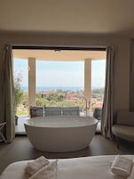 A modern bedroom with a large bathtub, a sofa, and a view of the city and sea.