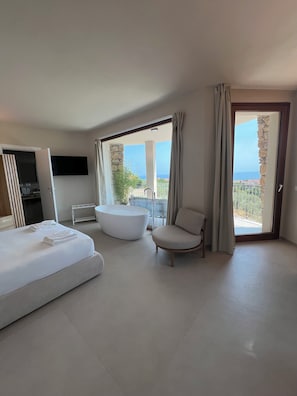 Habitación de hotel moderna con cama, bañera, sillón y balcón con vista al mar.