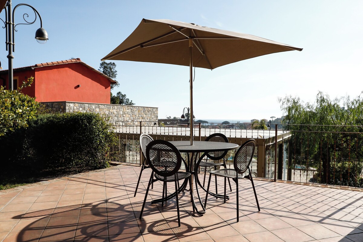An outdoor patio with a table and chairs, a large umbrella, and a view of the sea.