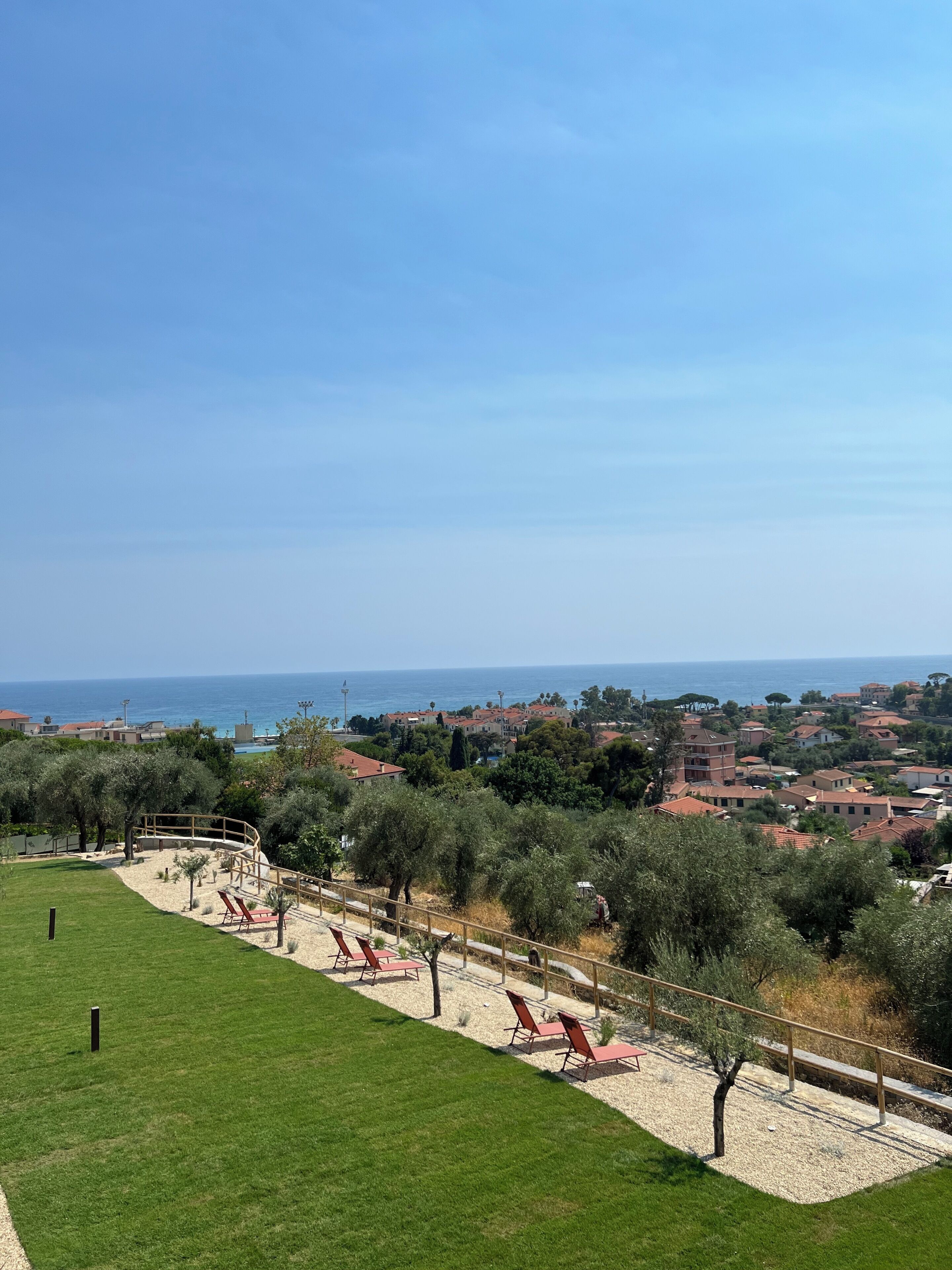 A coastal view with a grassy area, red lounge chairs, and a clear sky.