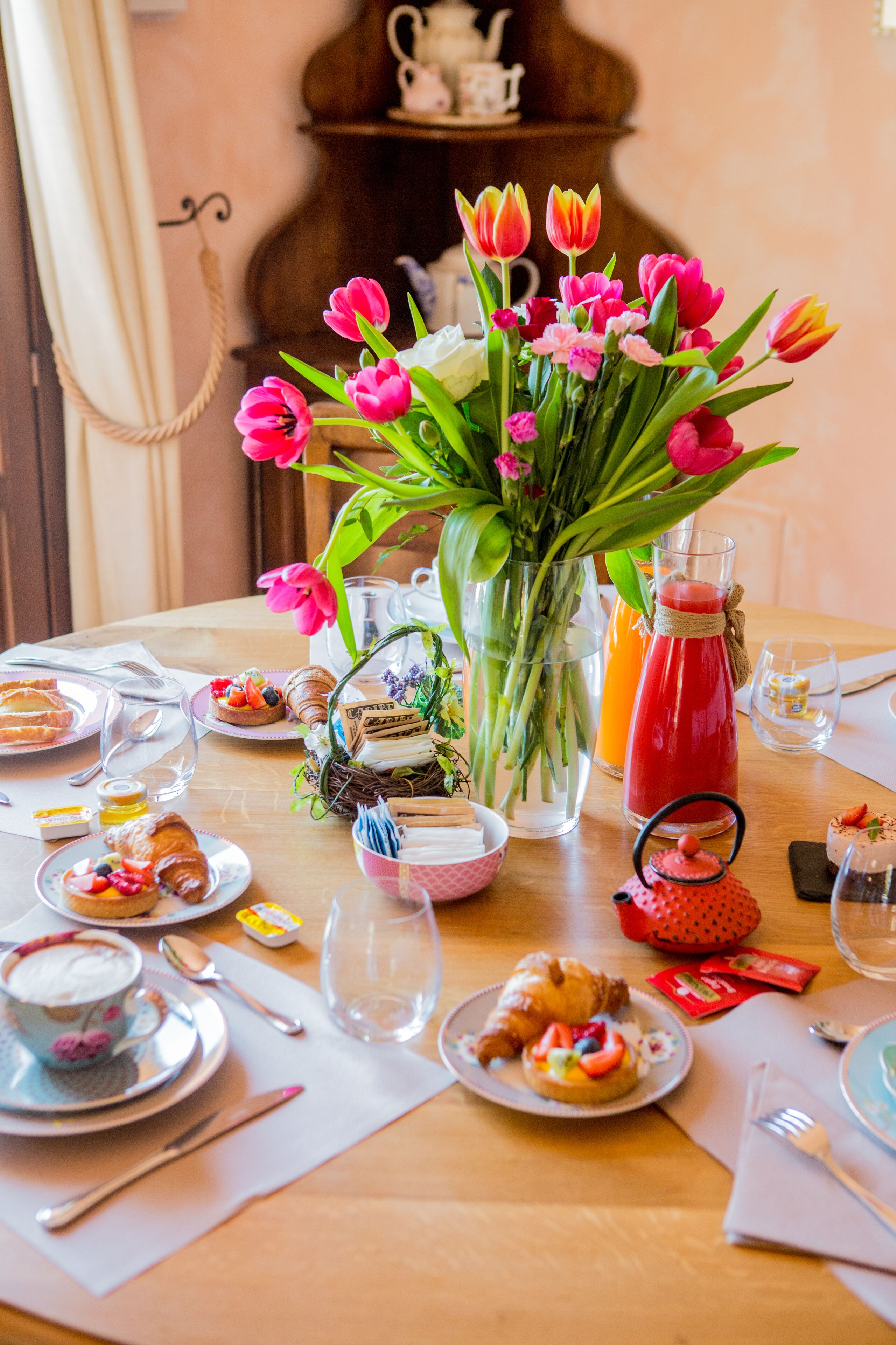 Una mesa de desayuno con varios platos, un jarrón de tulipanes y una jofaina de jugo.