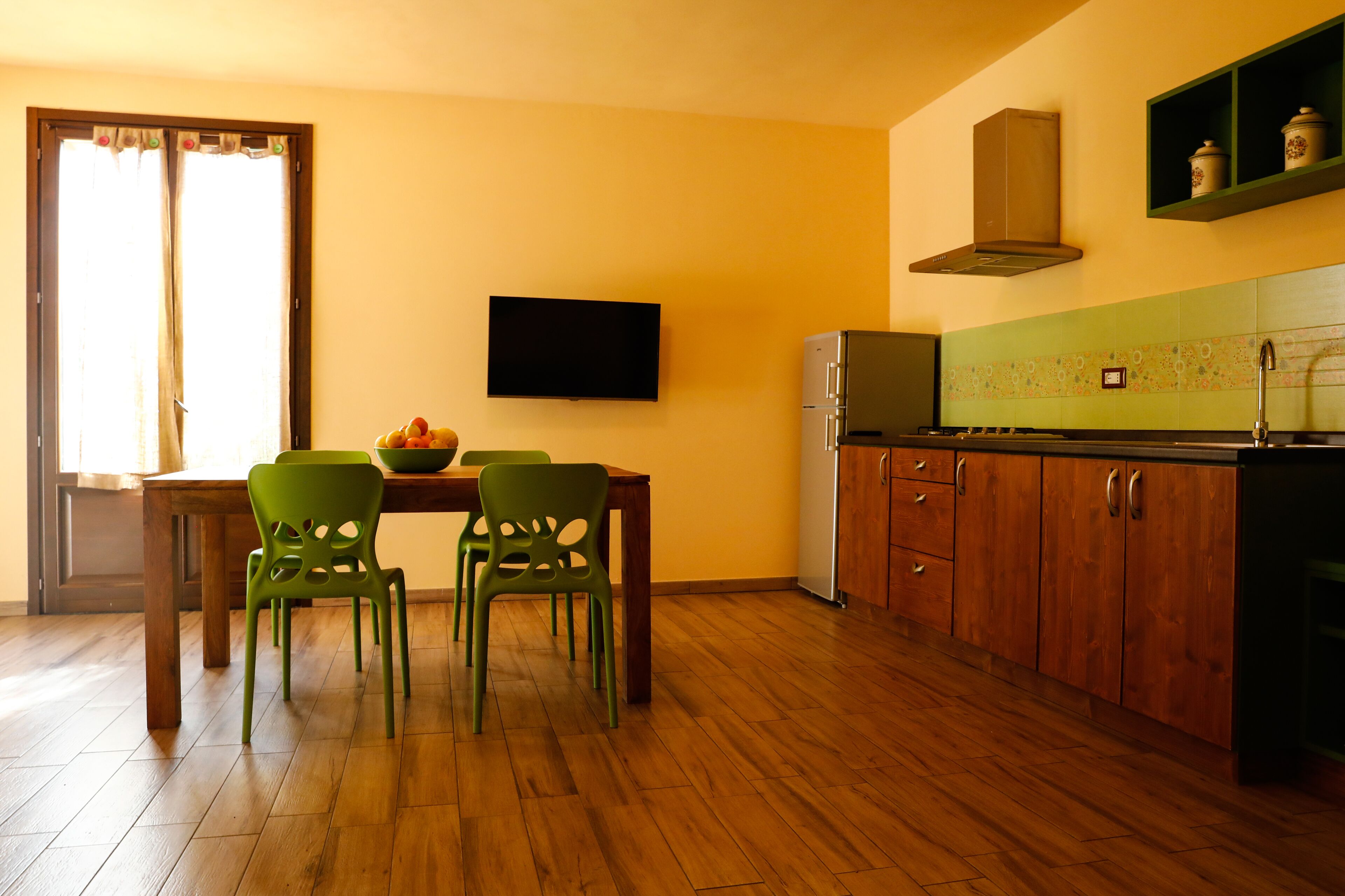 A kitchen with wooden cabinets, a refrigerator, a dining table with chairs, and a television mounted on the wall.
