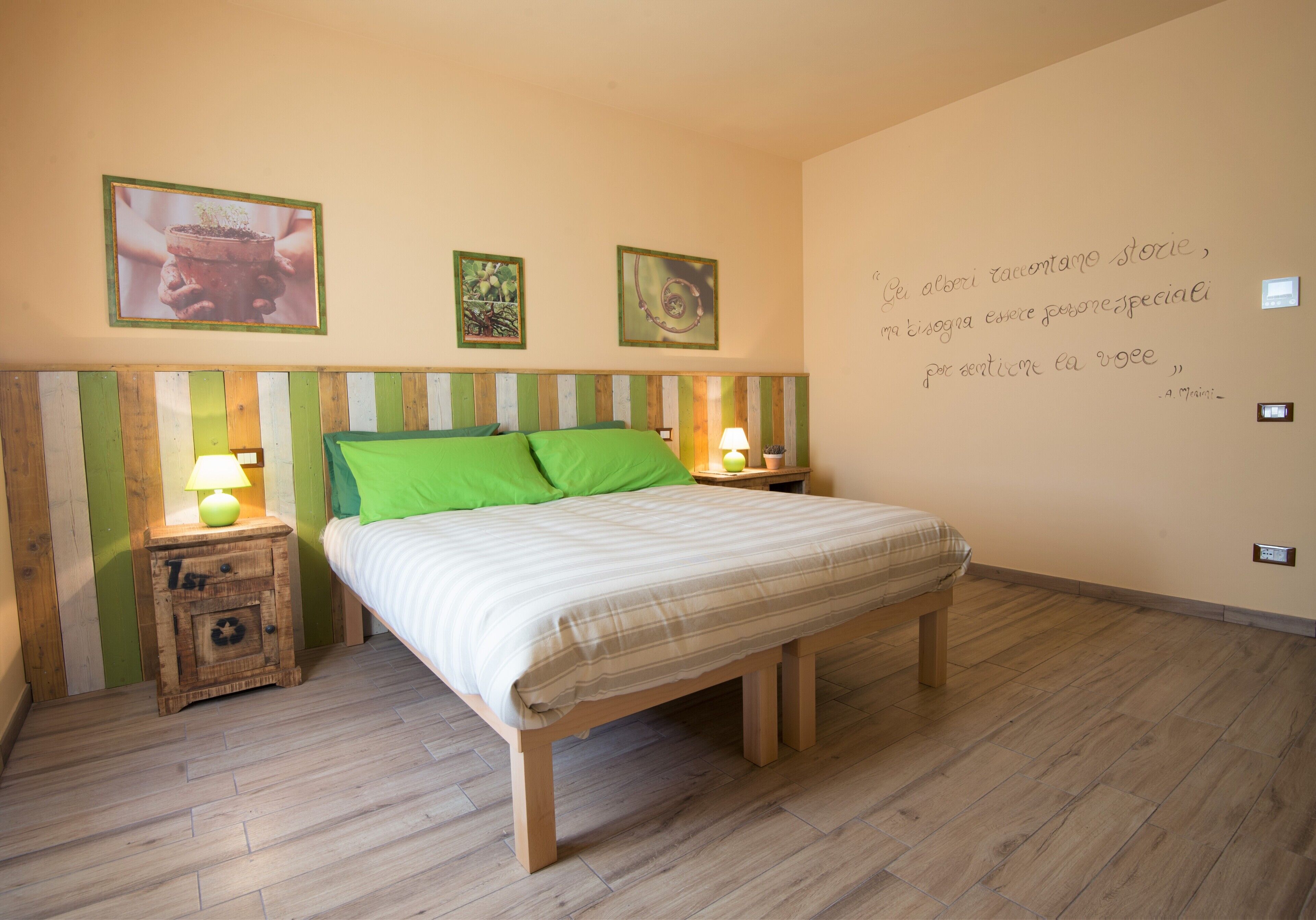 Een slaapkamer met een houten bed, groene kussens, een nachtkastje met een lamp, en een muur met ingelijste foto’s en een citaat.