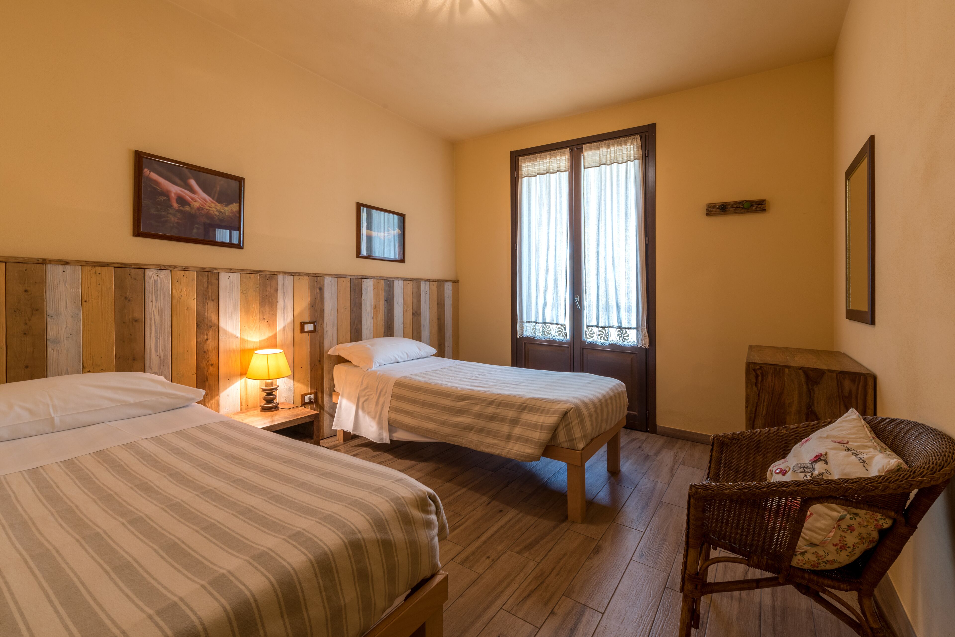 A room with two beds, a wooden headboard, a nightstand with a lamp, a wicker chair, and a window with curtains.