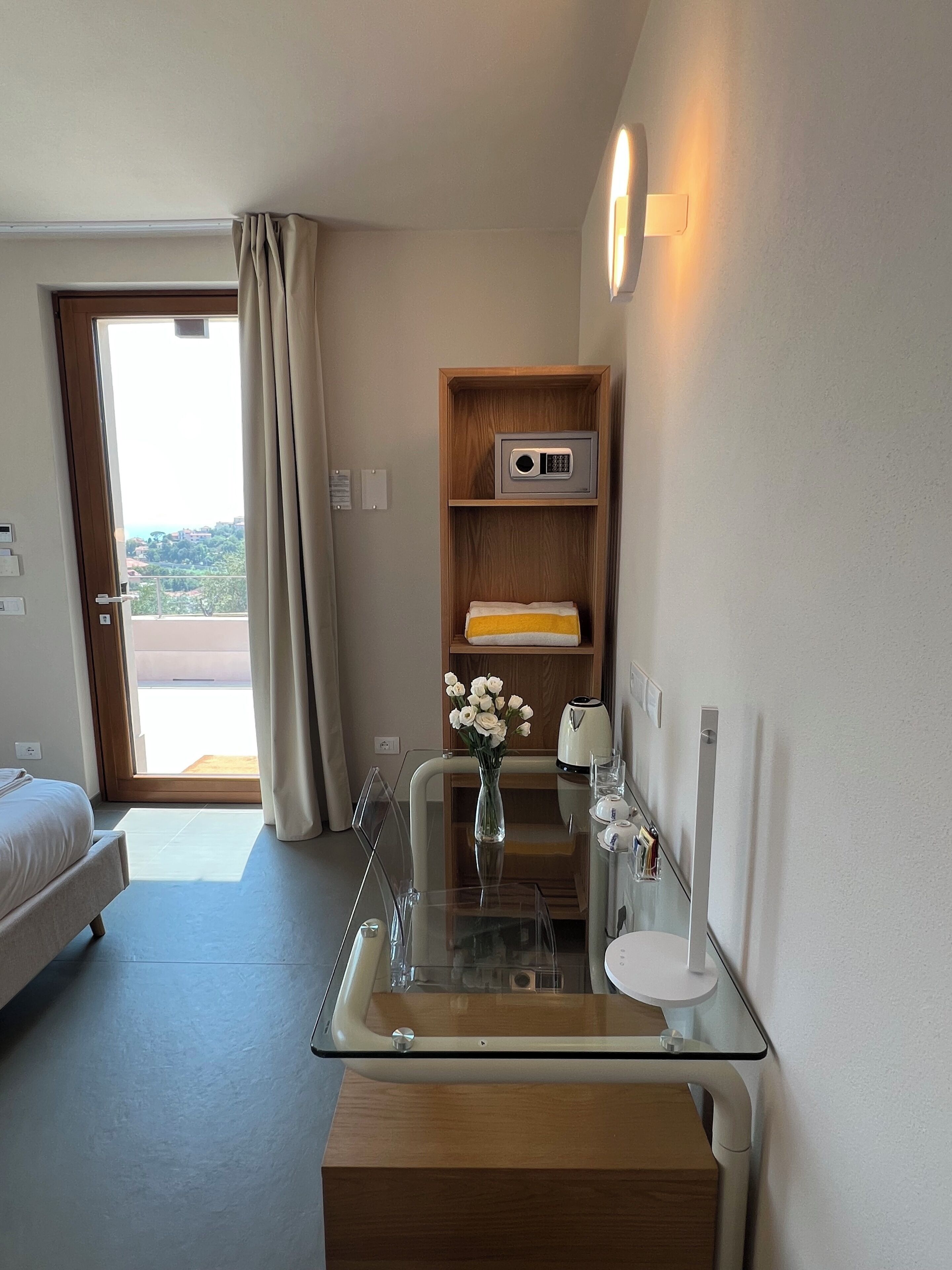 Ein Hotelzimmer mit einem Bett, einem Glastisch mit Blumenvase, einer Holzablage mit Wasserkocher und Blick durch eine Tür.