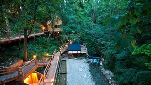 Un puente de madera que conecta un edificio con la orilla de un río, rodeado de un bosque frondoso.
