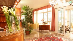 A hotel lobby with a reception desk, a large potted plant, a traditional rug, and a chandelier. - Hotel Abbazia (Grado)