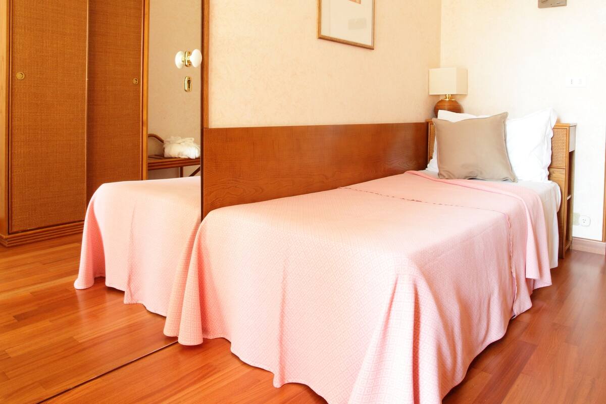 A hotel room with two beds, wooden flooring, a large mirror, and a framed picture on the wall.