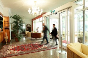 Un hall d’entrée avec un grand lustre, un tapis rouge à motifs, une plante en pot et un panneau indiquant la sortie.