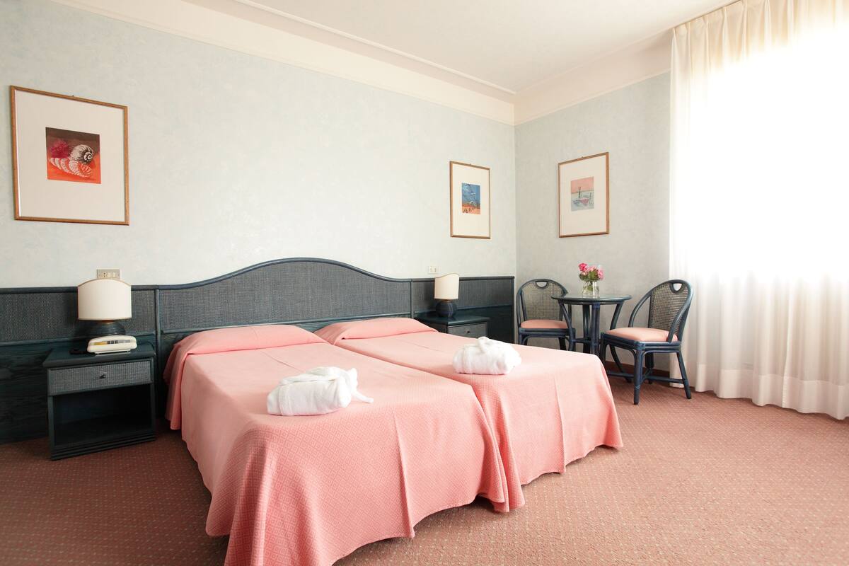 A hotel room with two beds, a nightstand with a lamp, a small table with a vase of flowers, and two framed pictures on the wall.