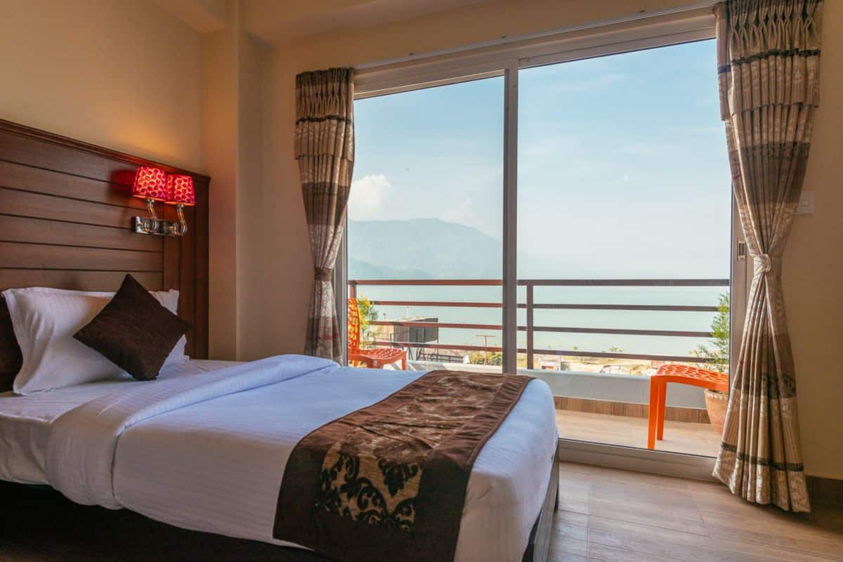 A hotel room with a large bed, a nightstand with a lamp, a view of the sea through a balcony, and a wooden headboard.