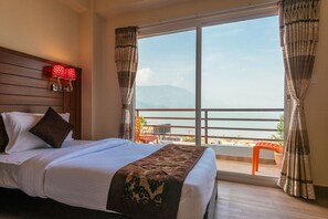 A hotel room with a large bed, a nightstand with a lamp, a view of the sea through a balcony, and a wooden headboard. - Hotel The Coast (Pokhara)
