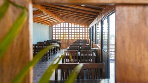 A restaurant with wooden tables and chairs, a high ceiling with exposed beams, and large windows allowing natural light. - Hotel The Coast (Pokhara)