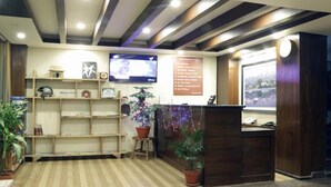 A reception area with a wooden reception desk, a television mounted on the wall, and a bulletin board with information. There are potted plants and a striped floor. - Hotel The Coast (Pokhara)