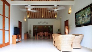 A spacious living area with a ceiling fan, a television, wicker furniture, and a dining table with chairs. - Villa Sutra (Pemuteran)