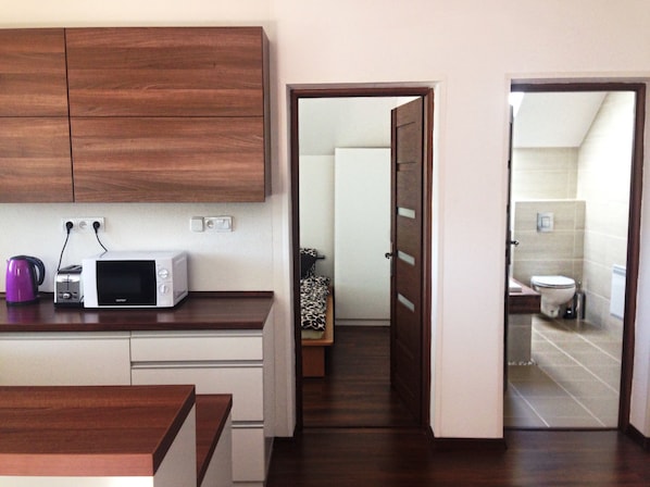 A kitchen with wooden cabinets, a microwave, and a toaster. There is a door leading to a bathroom with a toilet and sink. - Apartmány STAR (Bobrovec)
