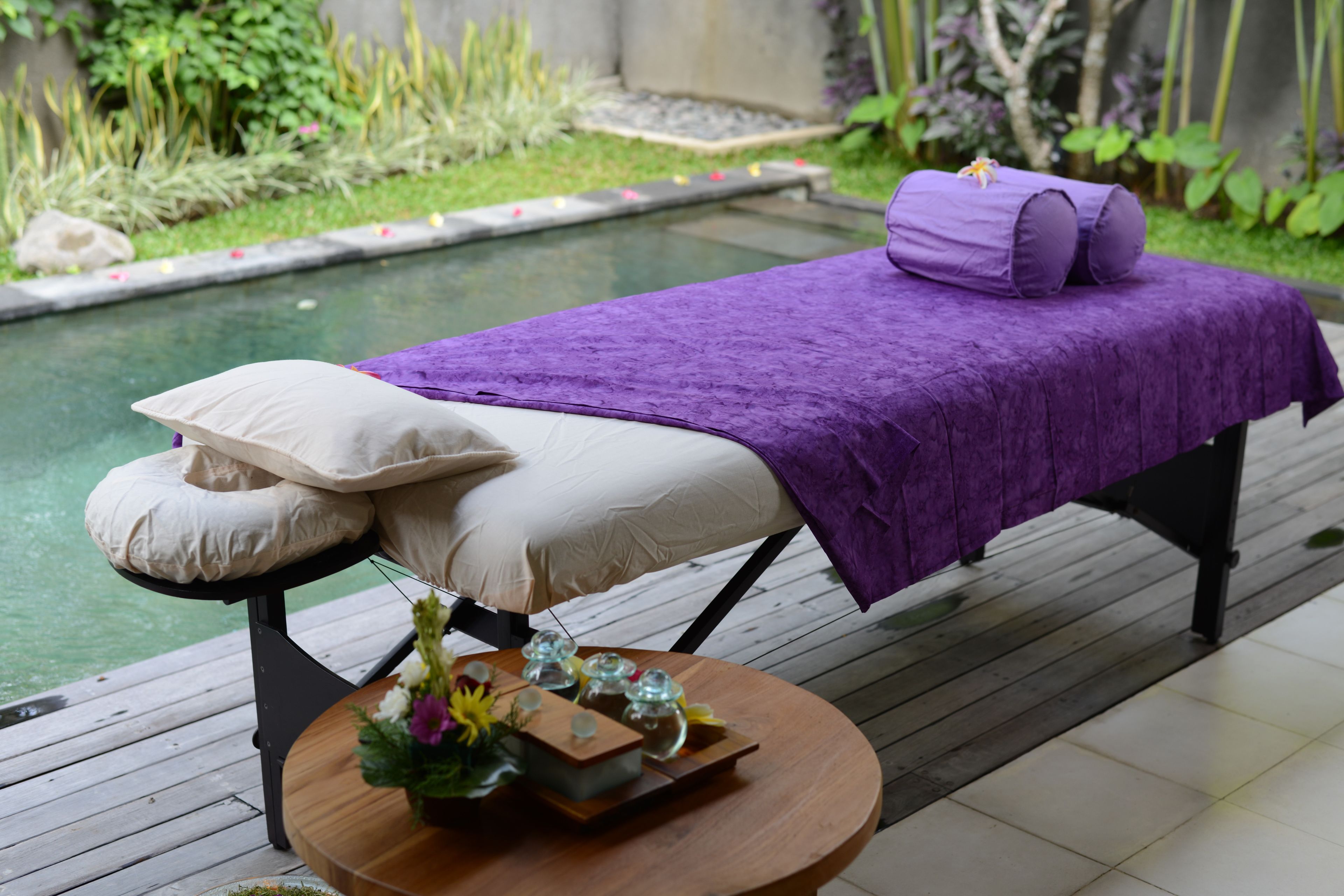 Una mesa para masajes con una toalla morada y almohadas, una mesa de madera con un florero y una piscina al fondo.