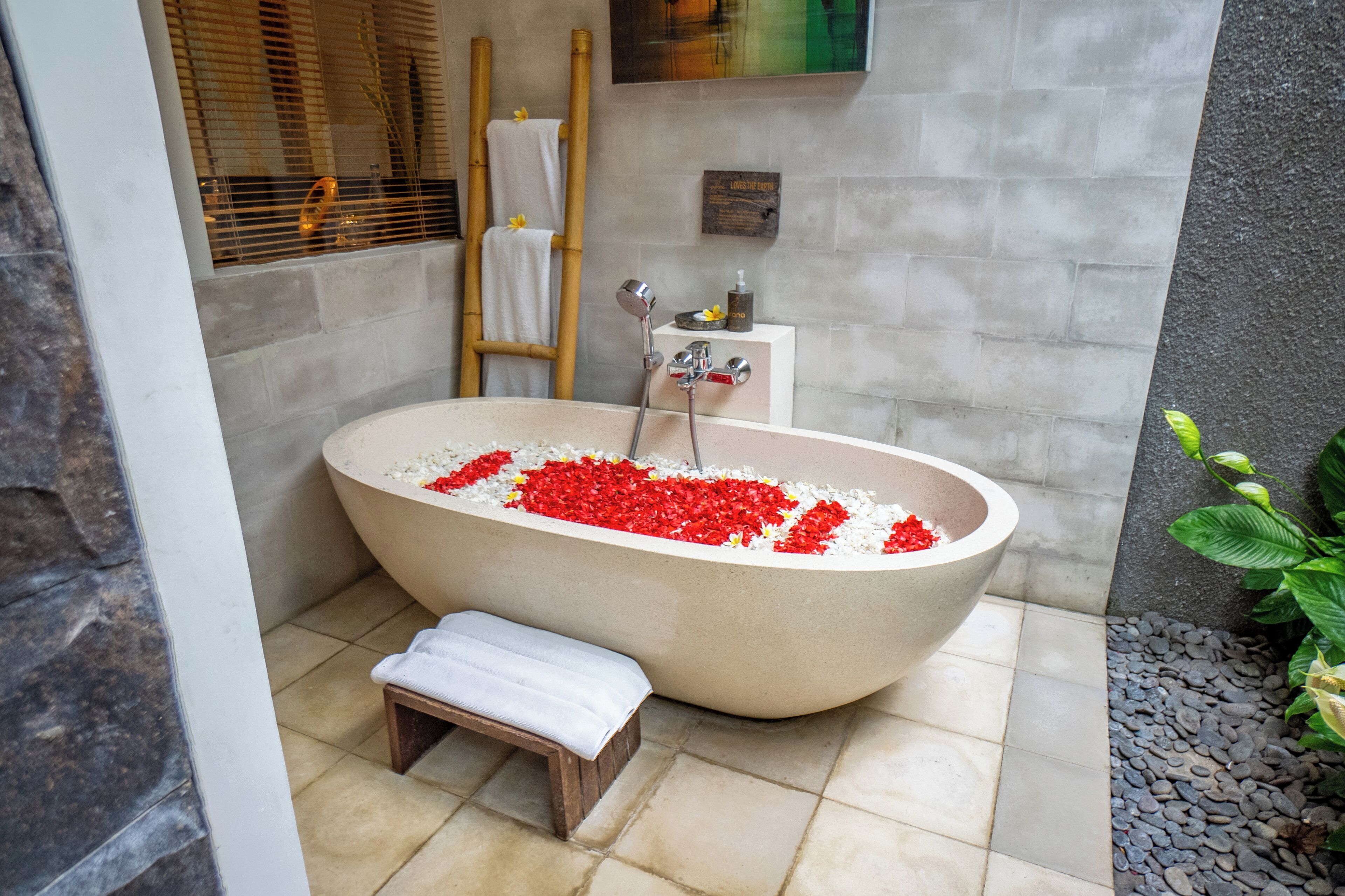 Un baño tipo spa con una bañera independiente llena de flores rojas, un banco de madera y una escalera de bambú.
