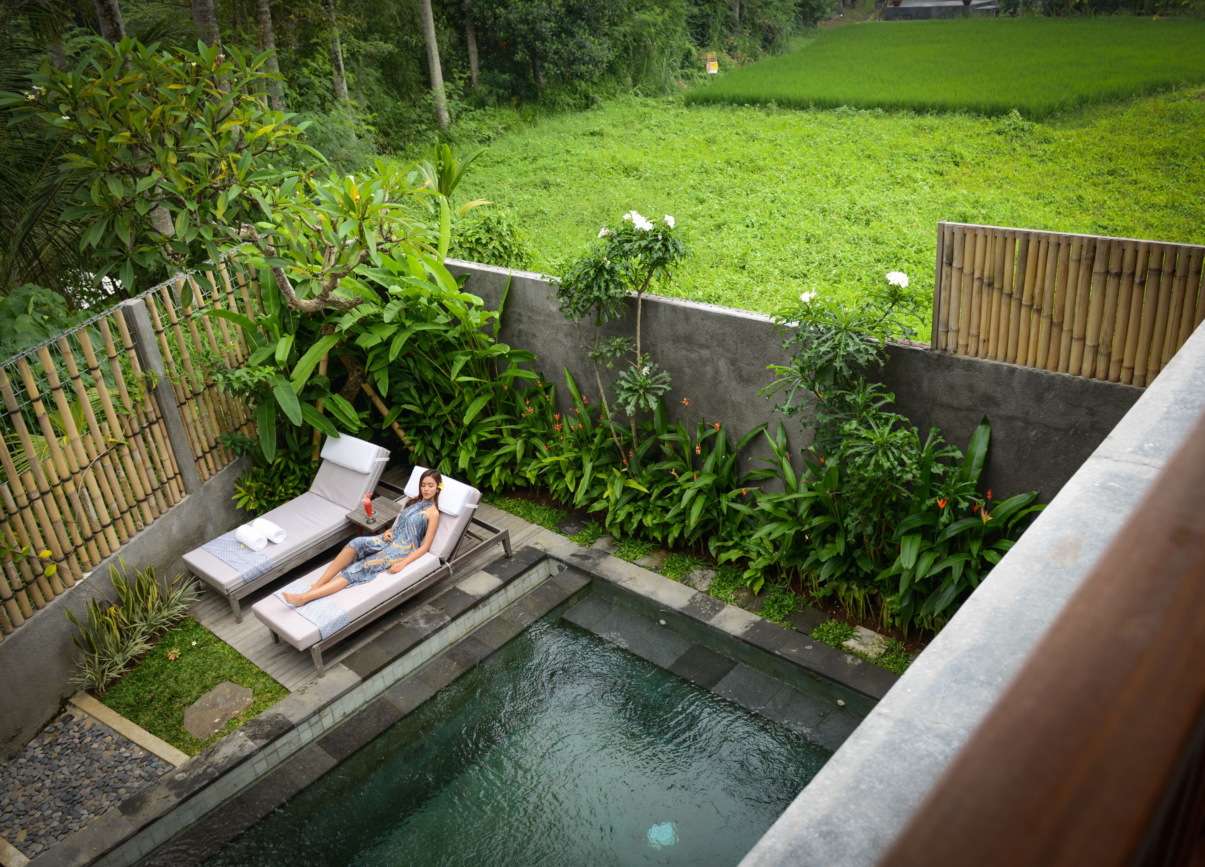 A pool area with two lounge chairs and a small table, surrounded by lush greenery and a bamboo fence.