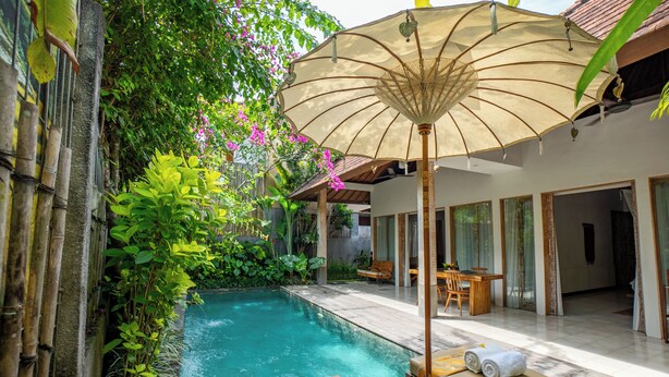 Zona de piscina con una silla de descanso, una mesa con toallas y un gran parasol.