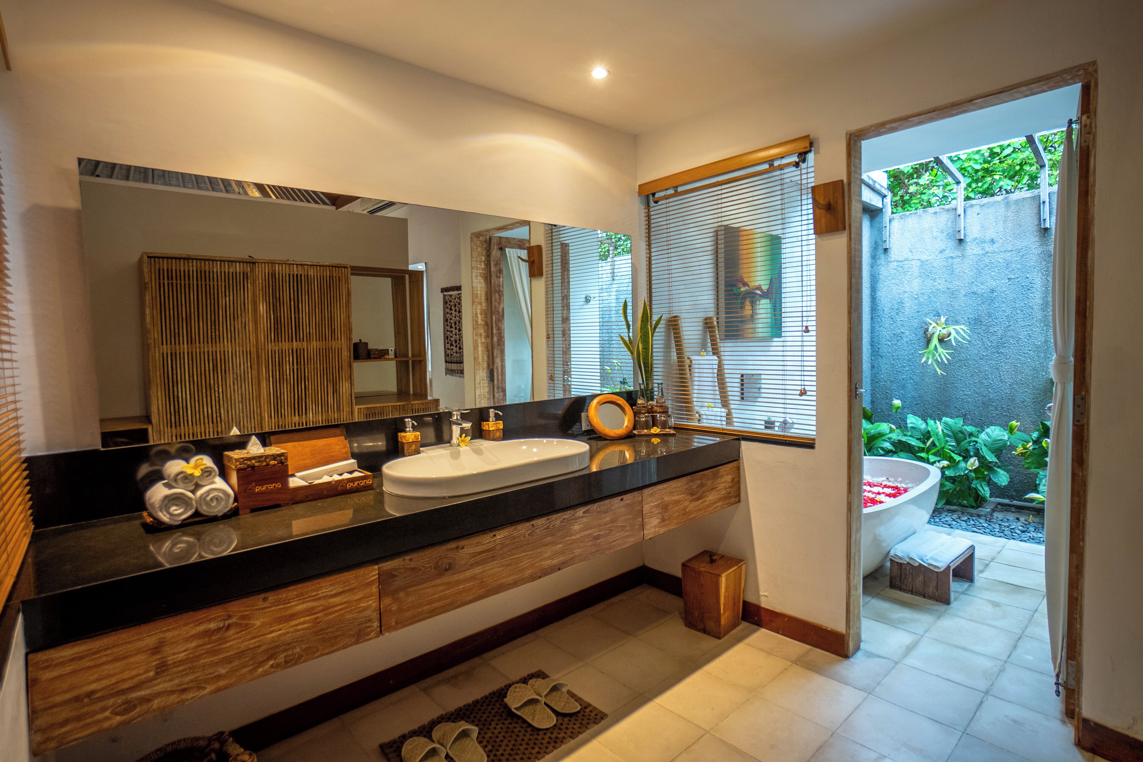 A modern bathroom with a large mirror, wooden vanity, dual sinks, and a view of a garden through the door.