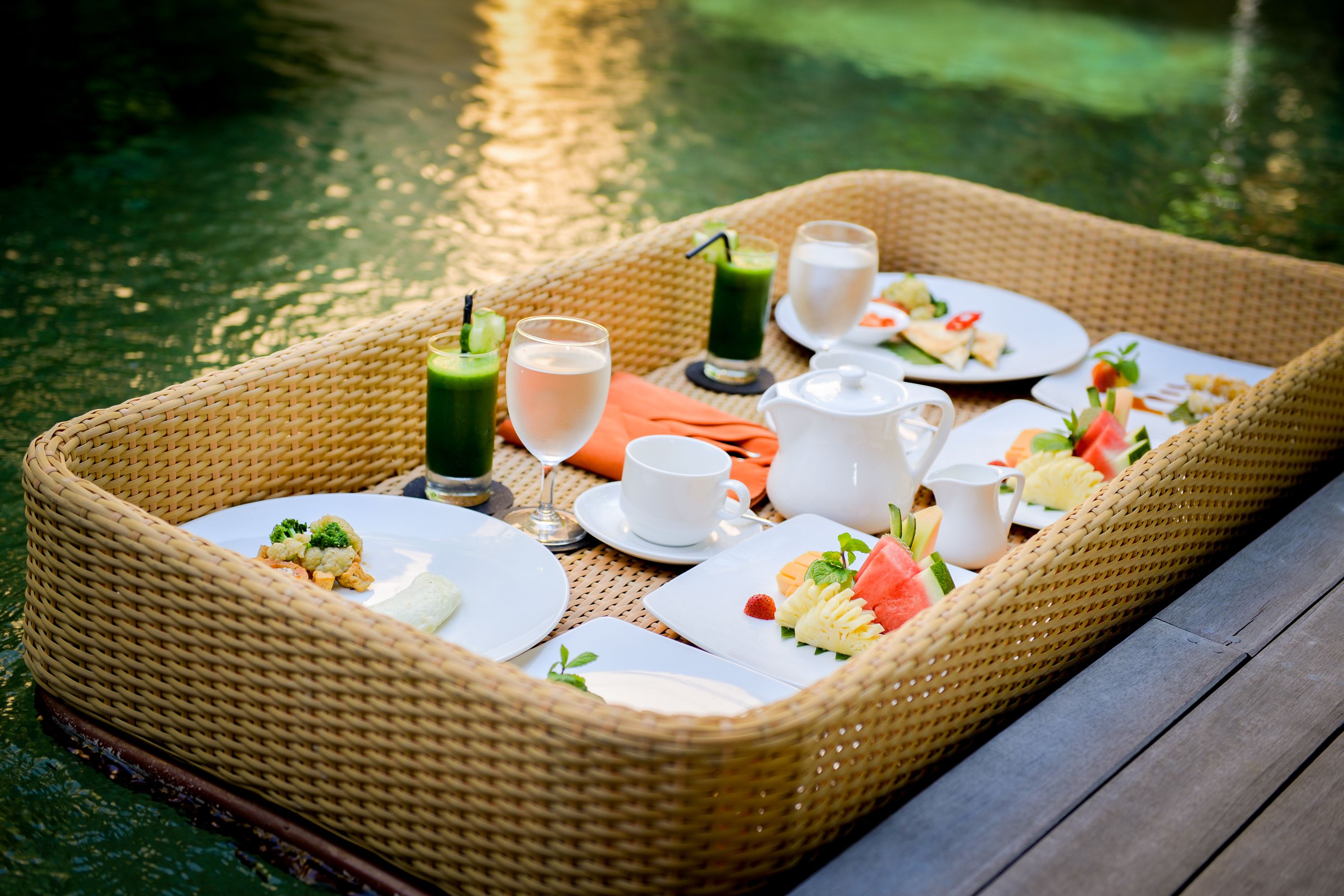 Een rieten dienblad met een picknickopstelling aan het water, inclusief borden met eten, drinken en een theepot.