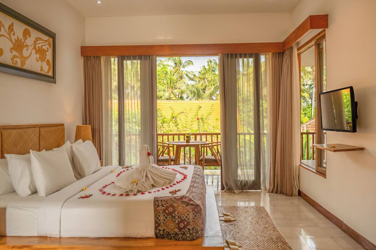 A hotel room with a bed, a TV, a balcony with curtains, and a view of greenery.