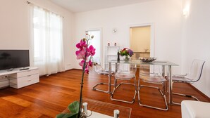 Uma sala de estar moderna com uma mesa de jantar de vidro, cadeiras transparentes, uma televisão de tela plana e uma orquídea em vaso.