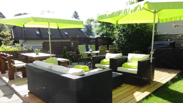An outdoor patio with wicker furniture, green cushions, and a wooden deck. - Pension AdlerHorst (Steindorf am Ossiacher See)