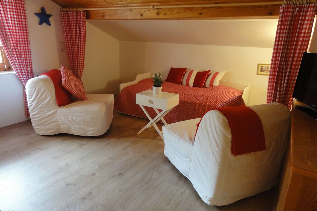 Une pièce chaleureuse avec deux fauteuils blancs, un lit à motifs rouges et blancs, et une petite table blanche sur laquelle est posée une plante.