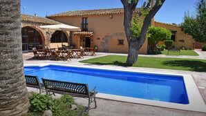 Una piscina con zona de patio, mobiliario exterior y un edificio con arcos y tejado de azulejos.