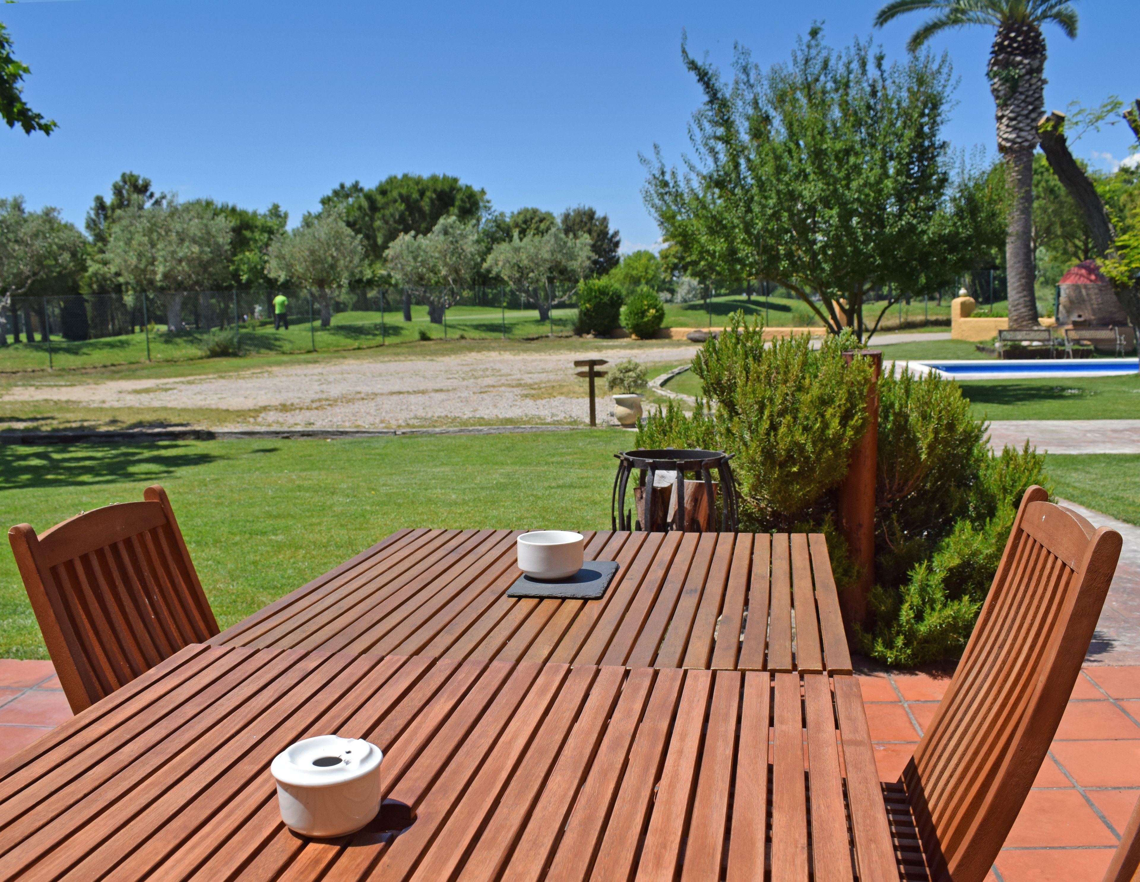 A wooden table with chairs, a bowl, and a cup on a patio with a view of a park and a pool.