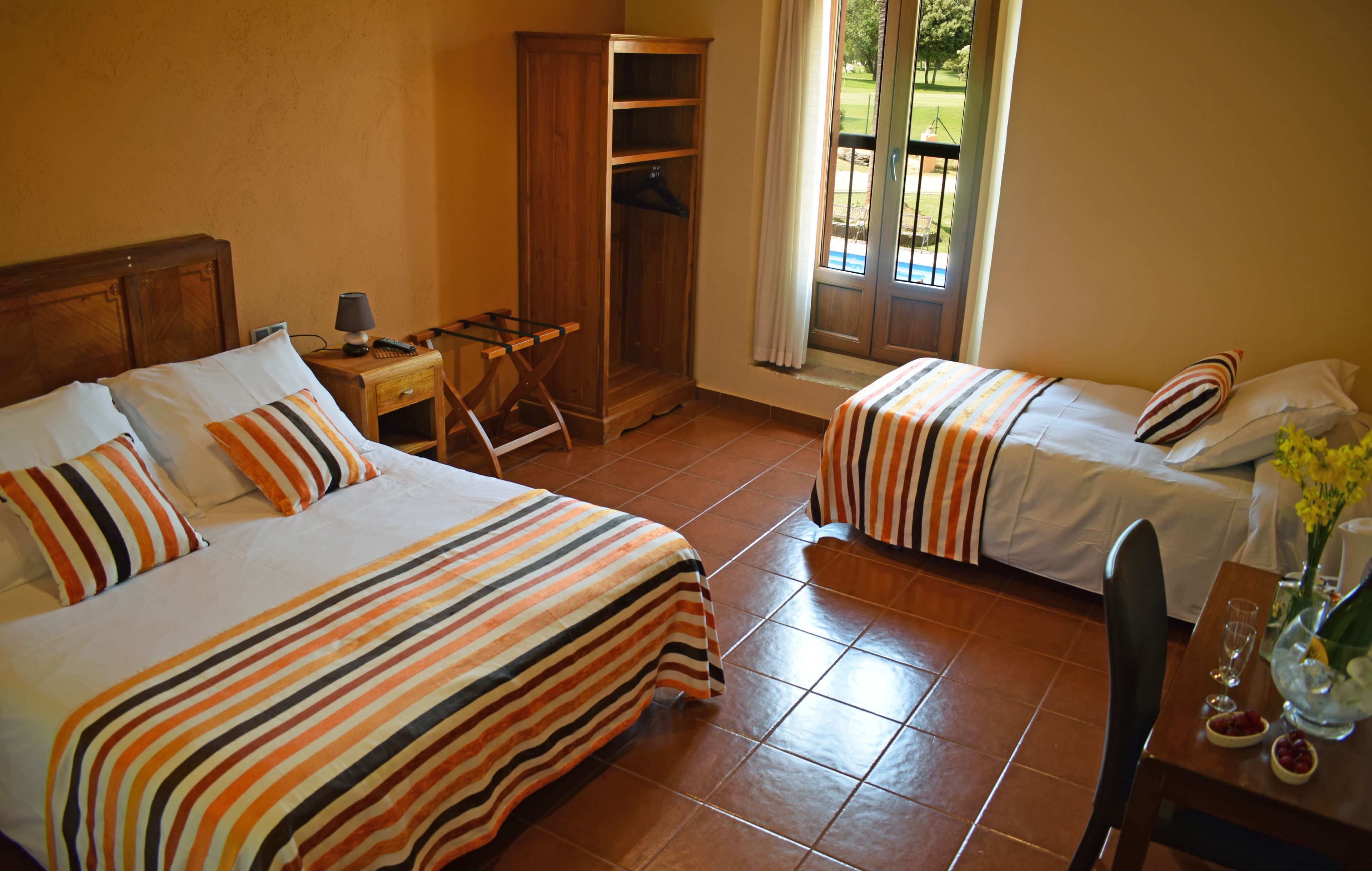 Quarto de hotel com duas camas, guarda-roupa de madeira, escrivaninha com abajur e vista para o exterior.
