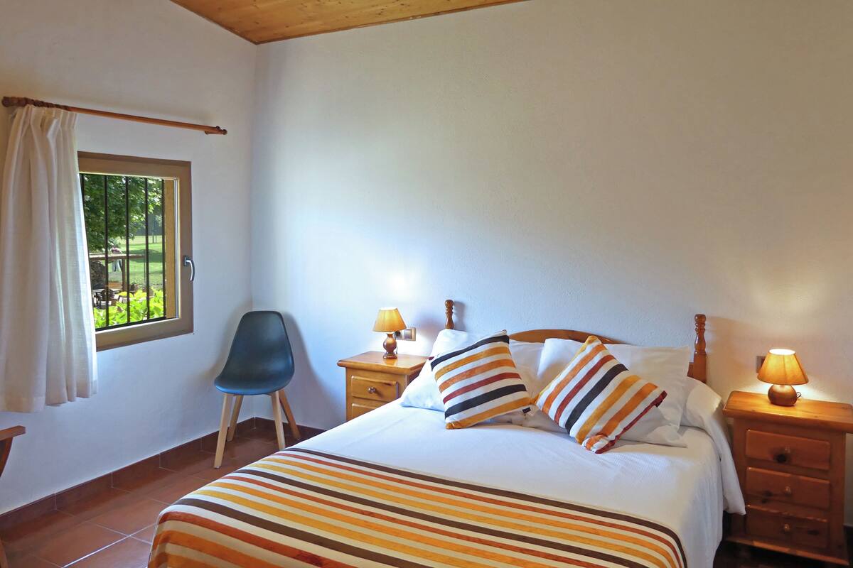 A bedroom with a bed, two bedside tables with lamps, a chair, a window with curtains, and a view of greenery outside.