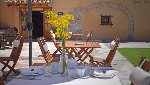 Un espacio exterior para comer con mesas y sillas de madera, un jarrón con flores amarillas y vistas a un edificio con un arco de piedra.