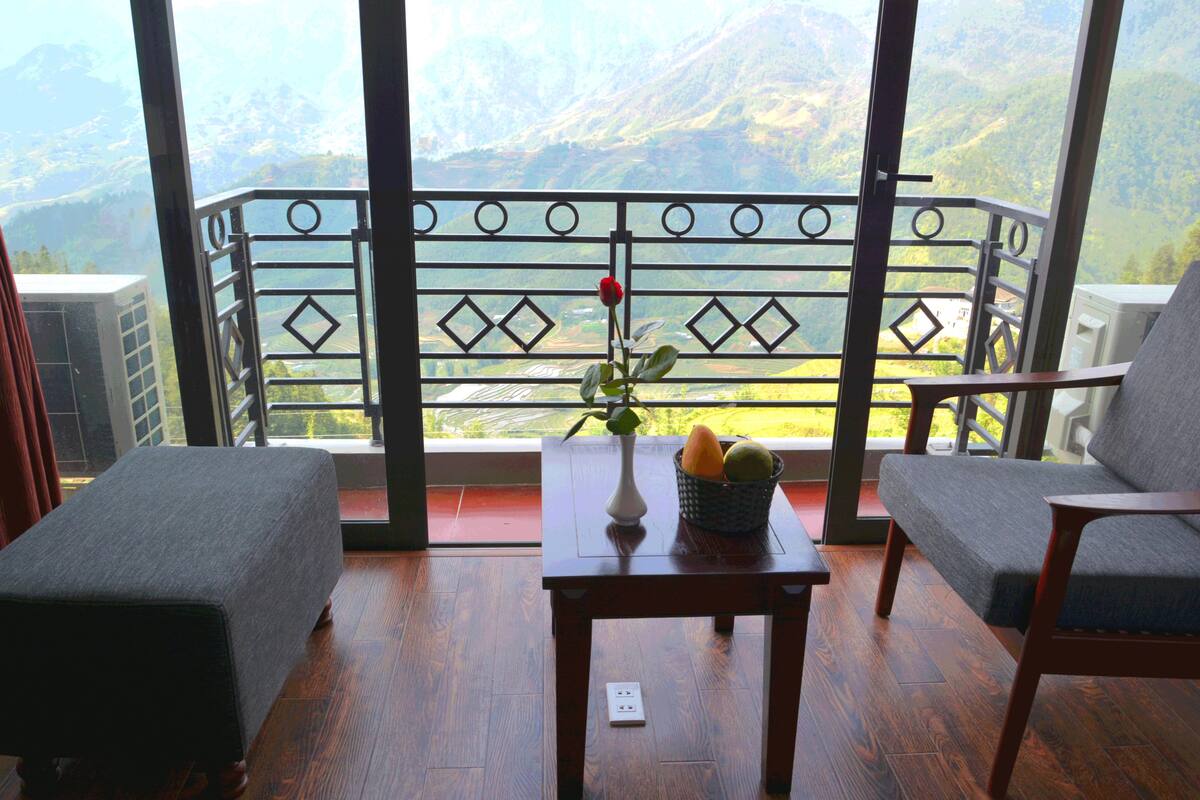Een balkon met uitzicht op bergen, een tafel met een vaas en een fruitmand, en twee gestoffeerde stoelen.
