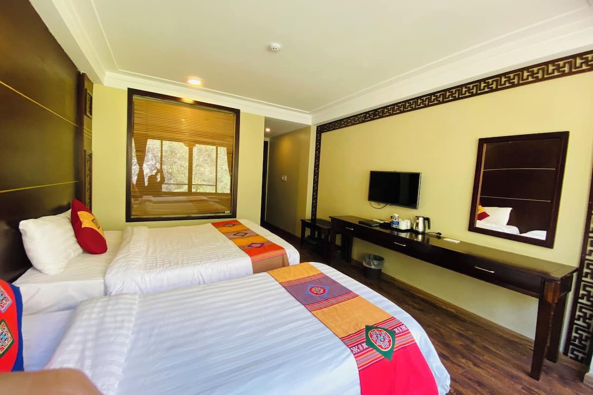 A hotel room with two beds, a TV, a desk, and a window with blinds.