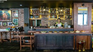 Een bar met een barman, houten tafels en een divers aanbod aan flessen en glazen op planken.