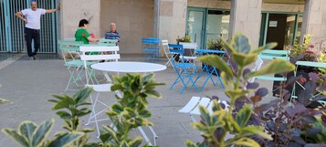 Zona all'aperto con tavoli e sedie, ingresso di un edificio e piante in vaso.