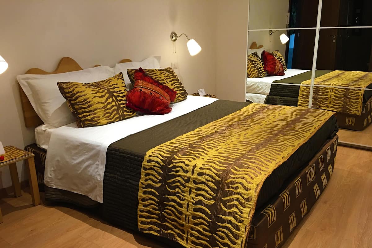 Una cama bien tendida con ropa de cama estampada, una mesita de noche con una lámpara y un espejo que refleja la habitación.