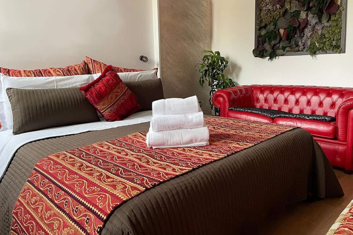 Habitación de hotel con cama, sofá de cuero rojo, colcha estampada y planta en la pared.