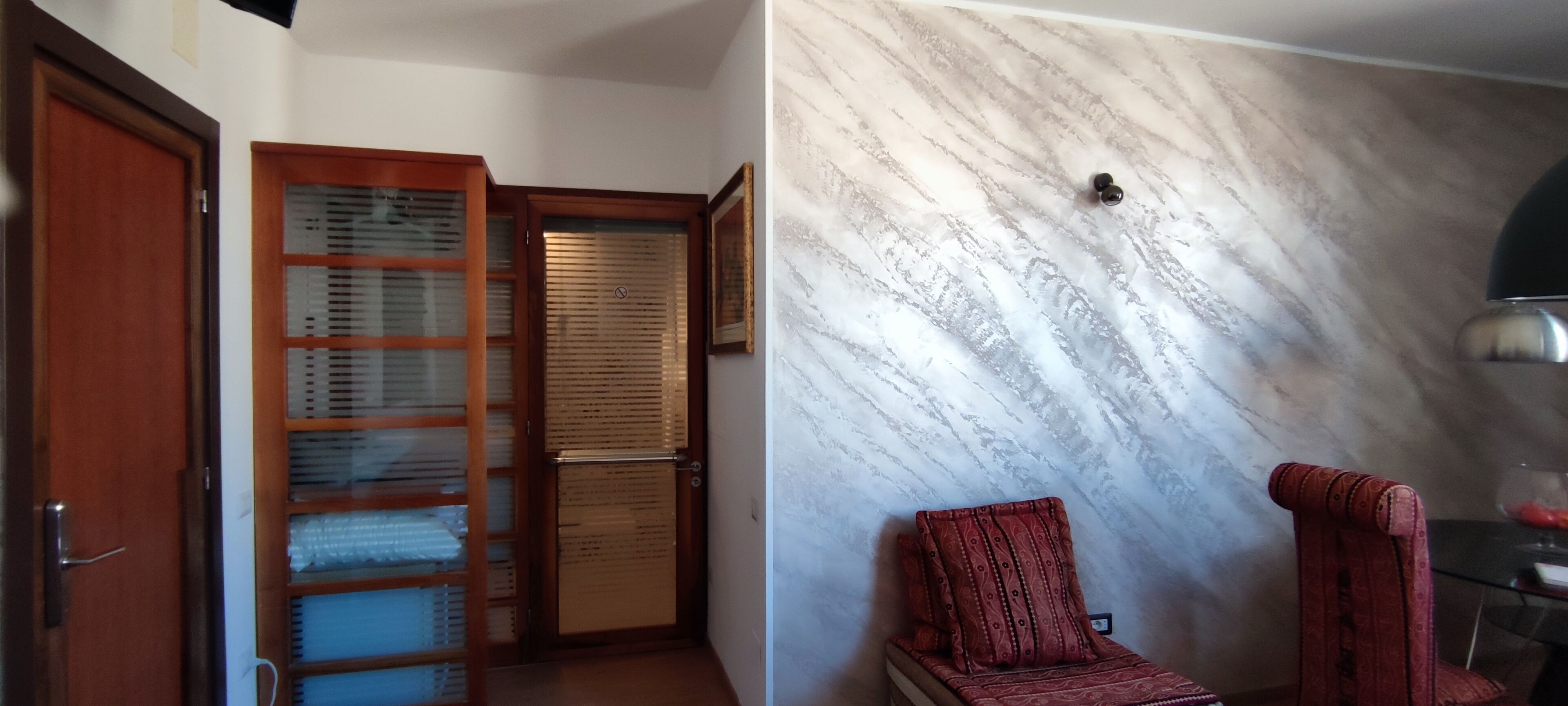 Ein Zimmer mit einer Holztür, einer Vitrine, einem rot gemusterten Sessel und einer Wand mit Marmoroptik.
