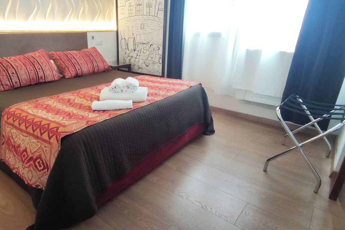 Una cama bien hecha con ropa de cama estampada, una mesita de noche, una lámpara de pared y una ventana con cortinas.
