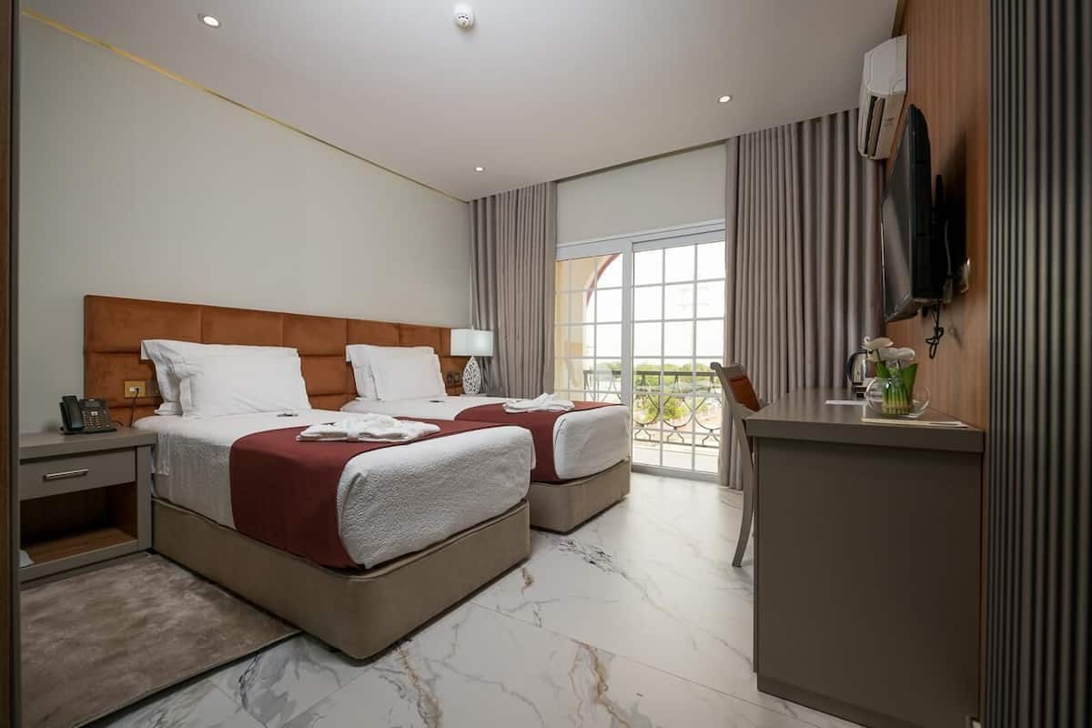 A hotel room with two beds, a desk, a TV, and a balcony with curtains.