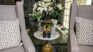 A seating area with two grey upholstered chairs, a round table with a green vase and flowers, and two champagne bottles. - Hotel Florença (Luanda)