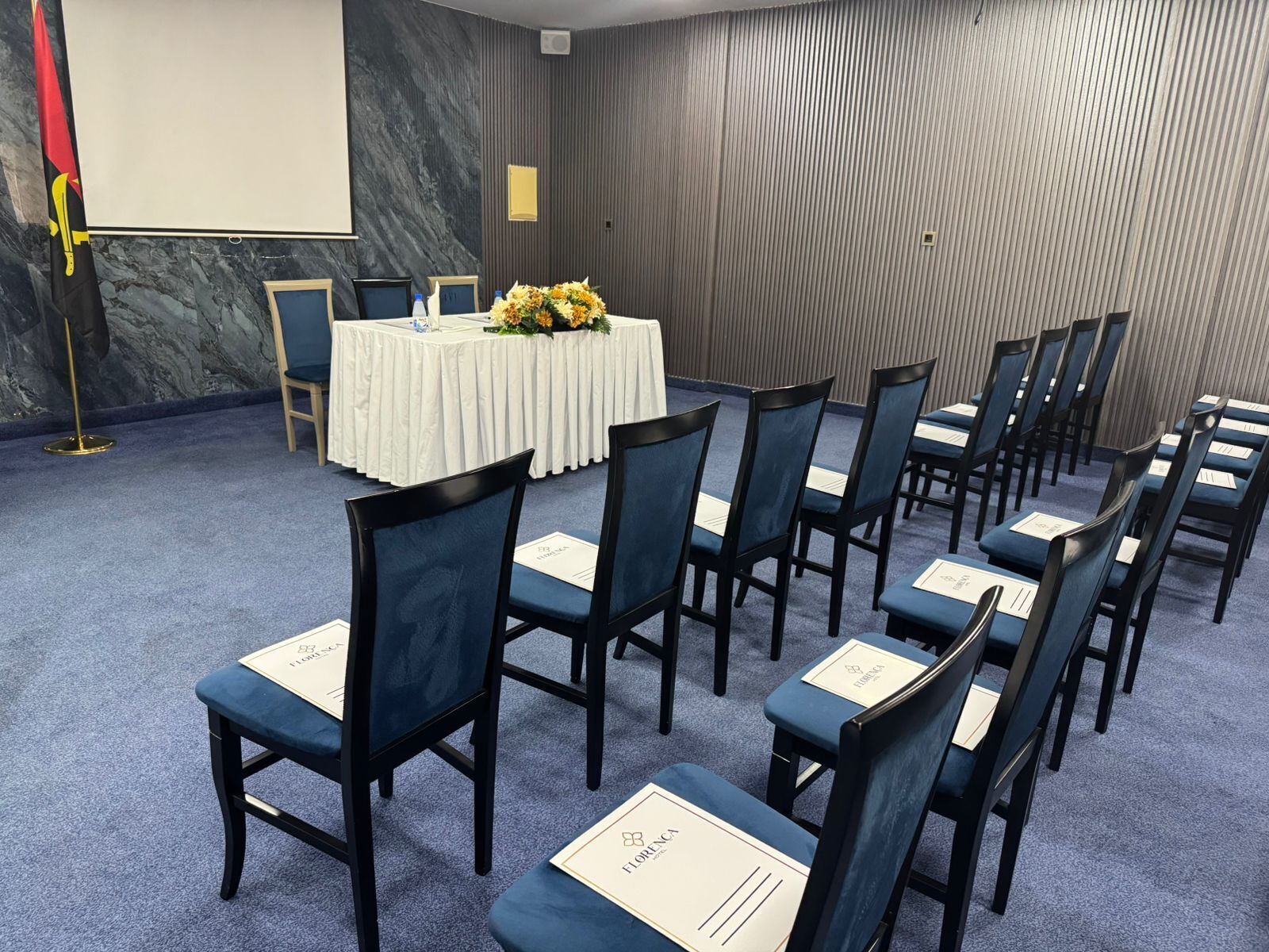 Konferenssihuone, jossa on riveissä tuoleja, valkoisella pöytäliinalla peitetty pöytä ja projektorikangas.