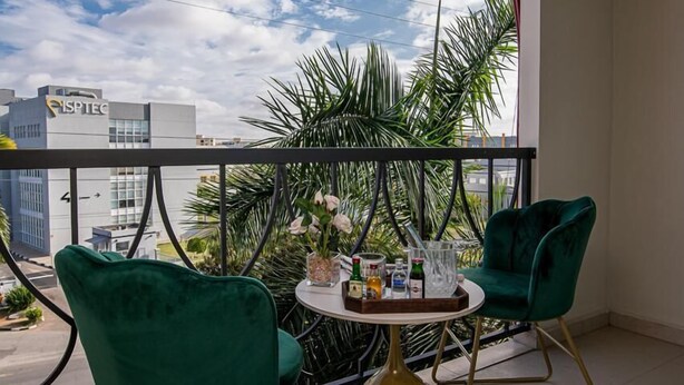 A balcony with two green velvet chairs, a small round table with a vase of flowers, and a view of palm trees and buildings. - Hotel Florença (Luanda)