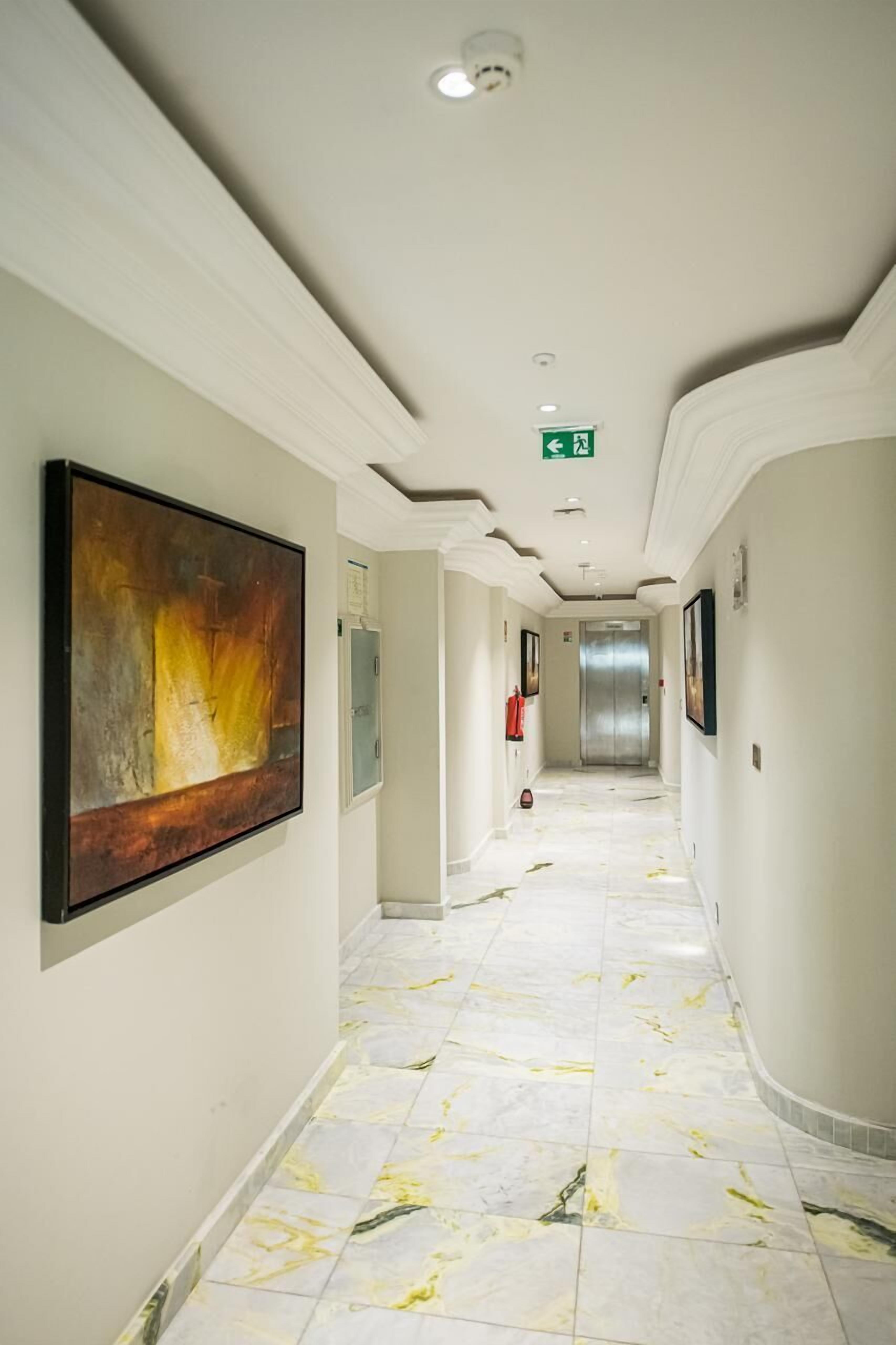 A hotel corridor with a marble floor, framed artwork on the walls, and an elevator in the distance.